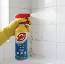 Dłoń w żółtej rękawicy trzymająca rozpylacz SAVO Przeciw pleśni na tle białych płytek łazienkowych.